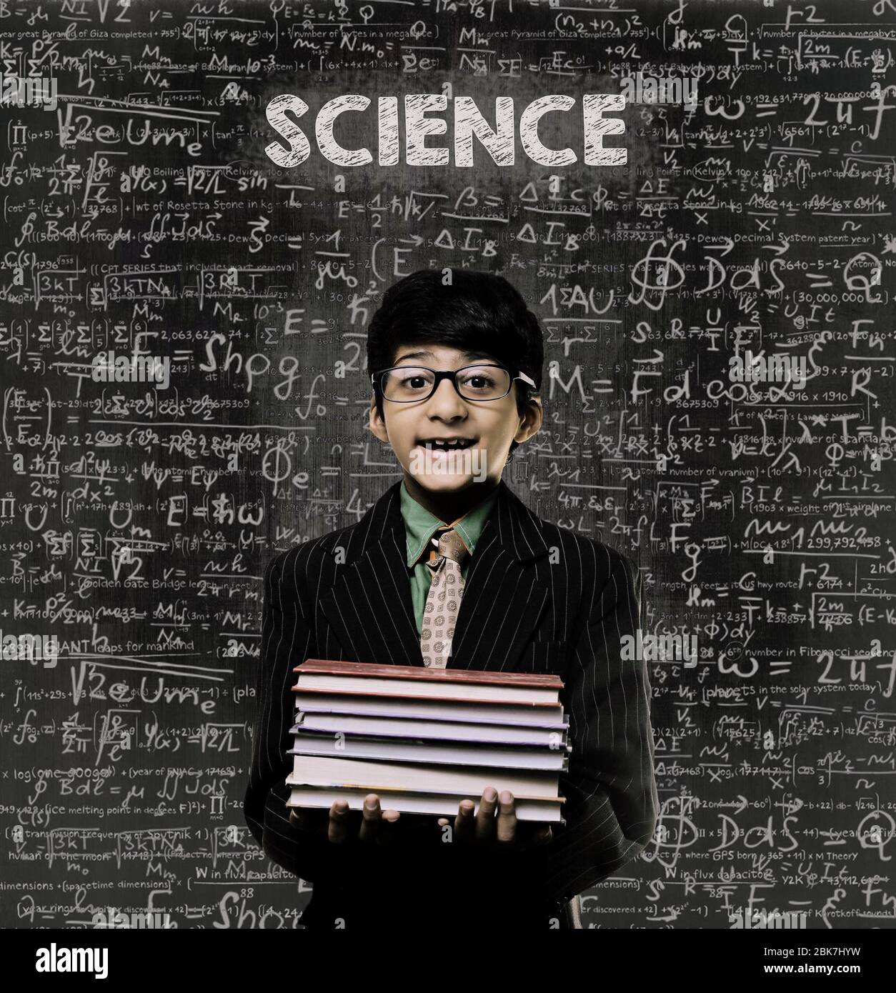 Cute Intelligent Little Boy Holding Book And Wearing Glasses Standing Before A Chalkboard, Chemical Formulas Are Written On Board With The Chalkboard Stock Photo