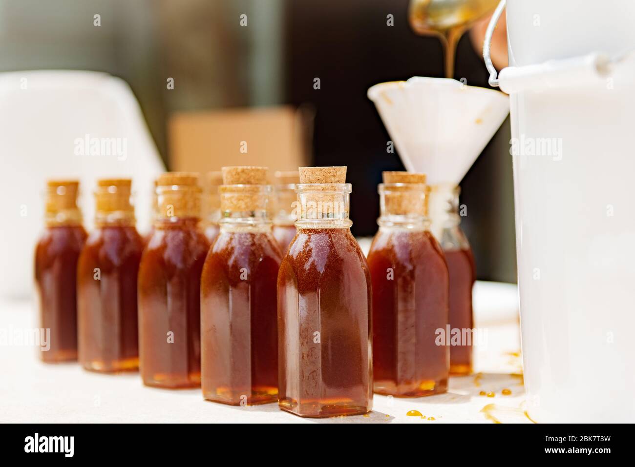 Honey in multiple 8 oz antique looking jars with cork lids and funnel in background, next to white bucket Stock Photo