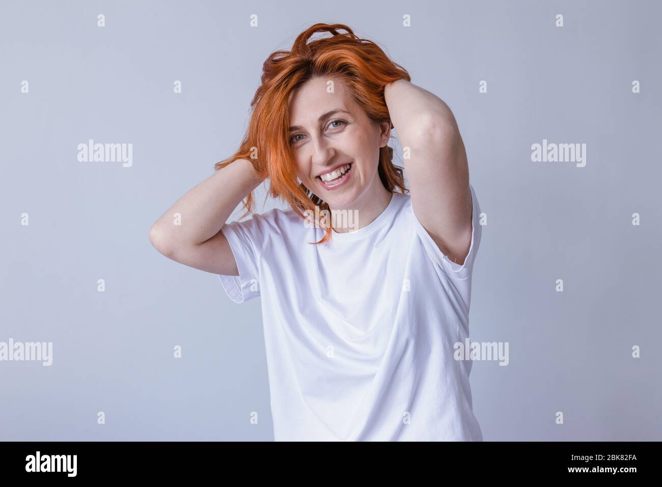 Horizontal portrait of pleasant-looking Caucasian female with long redhair,  wearing white casual T-shirt,  looking happily in camera Stock Photo