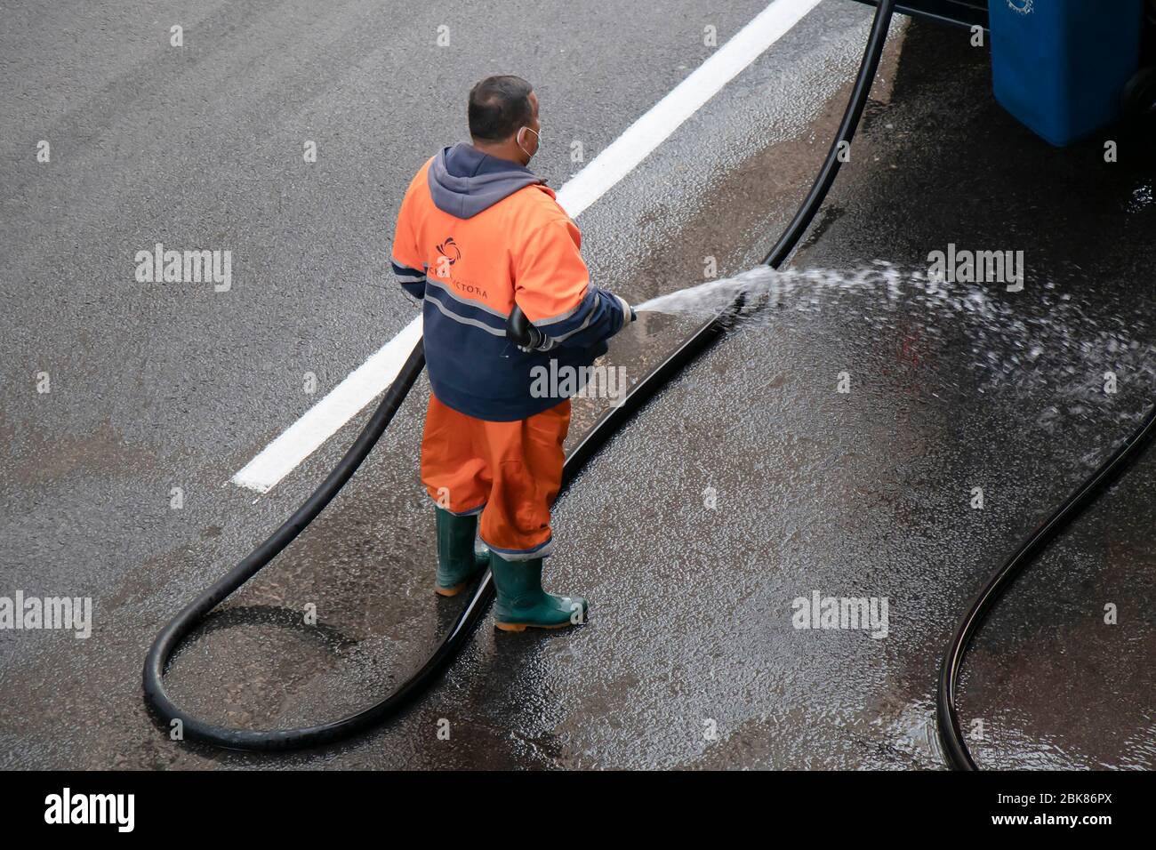 Belgrade, Serbia - April 24, 2020: Worker in orange uniform washing the street with water hose, high angle rear view; Stock Photo