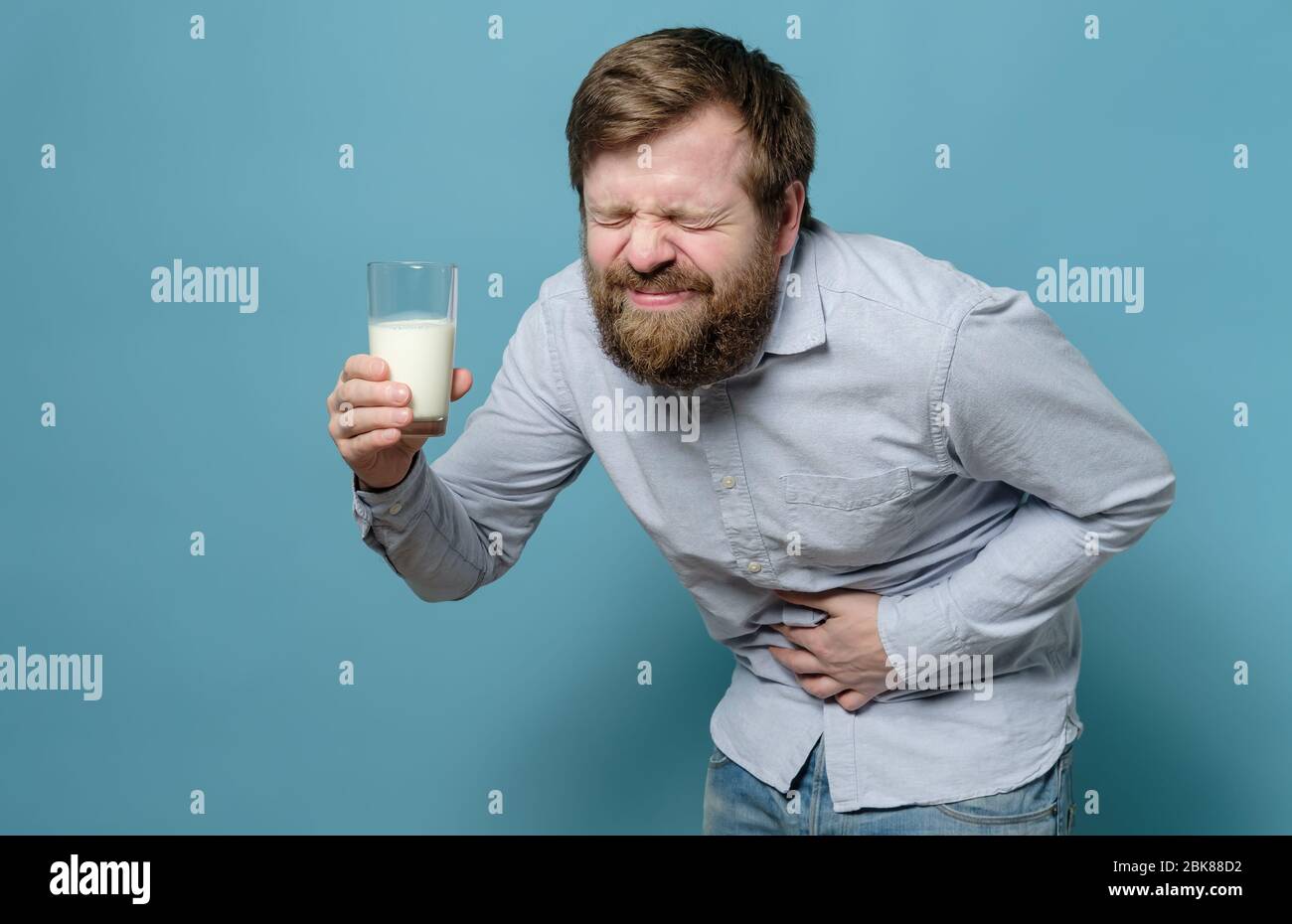 Lactose intolerance. Caucasian man holds a glass of milk in hand and suffers from severe pain in stomach, on a blue background. Stock Photo