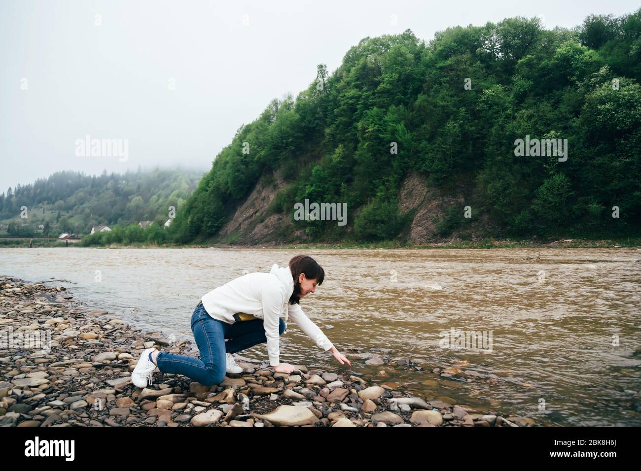 Tourist girl in jeans and white sweatshirt touching the yellow water on the bank of the dirty mountain river surrounded by forest Stock Photo
