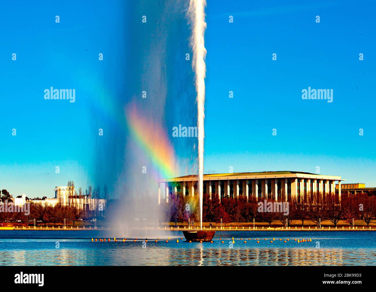 Rainbow through Captain Cook Memorial water jet on Lake Burley Griffin in Canberra, Australia's national capital, with National Library in background Stock Photo