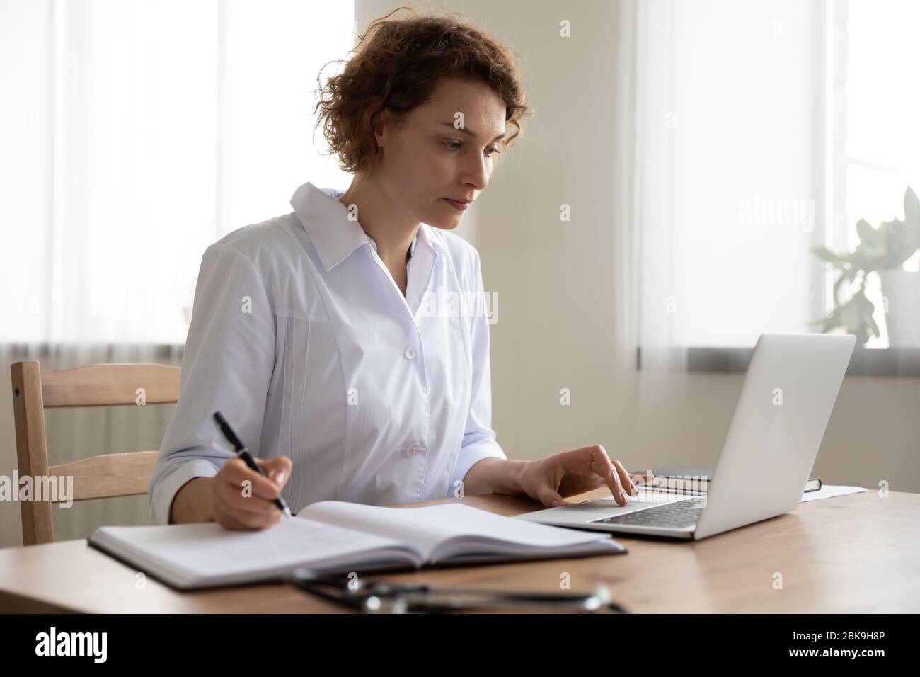 Young woman doctor working on laptop taking notes at workplace Stock Photo