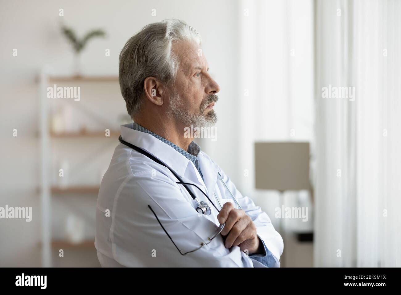 Thoughtful serious senior doctor looking through window lost in thoughts Stock Photo