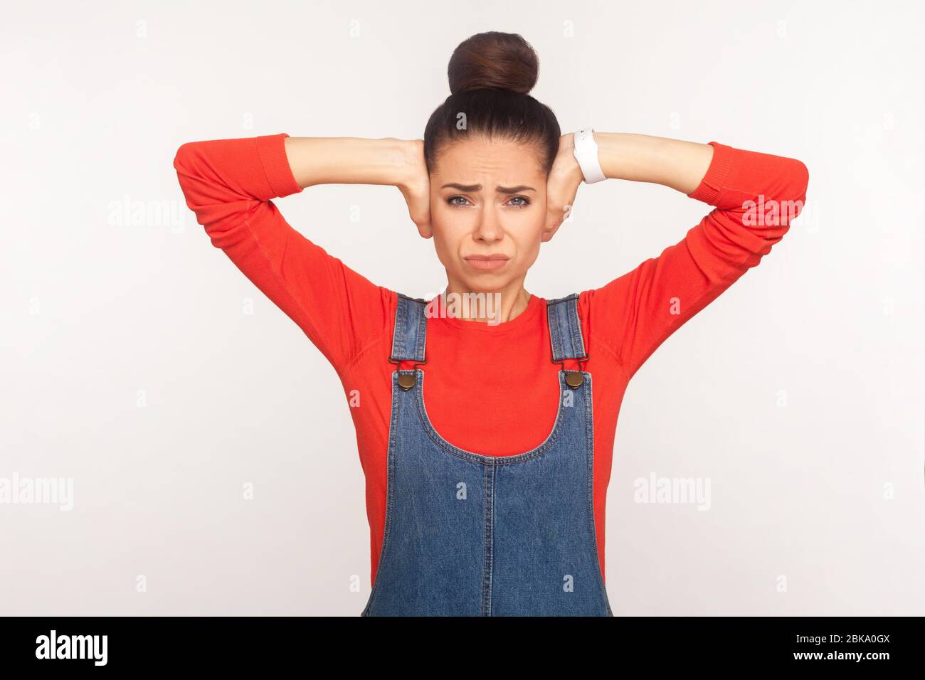 Don't want to hear! Portrait of irritated girl with hair bun in denim overalls desperately closing her ears from loud noise or bang sound, ignoring an Stock Photo