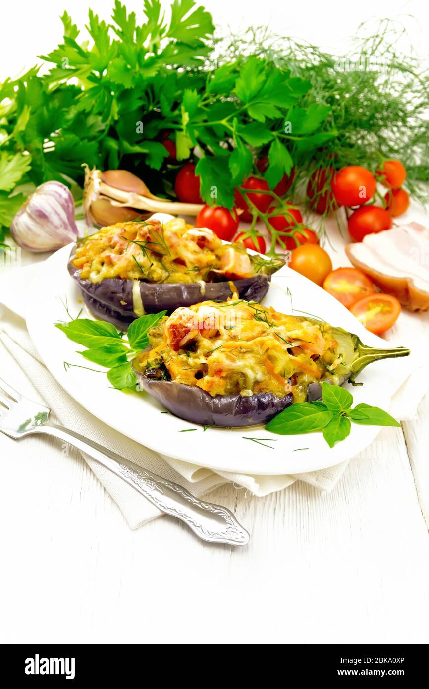 Stuffed eggplant with smoked brisket, tomatoes, onions, carrots with garlic, cheese and herbs in an oval plate on a napkin on background of light wood Stock Photo