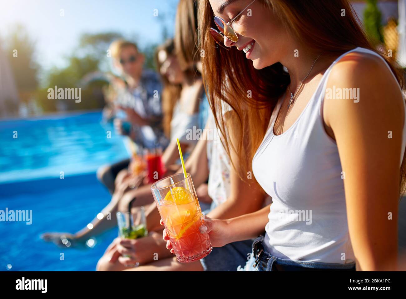 Pretty woman relax with fresh colorful cocktail glass sitting by swimming pool on sunny summer day with friends. People toast drinking beverages at Stock Photo