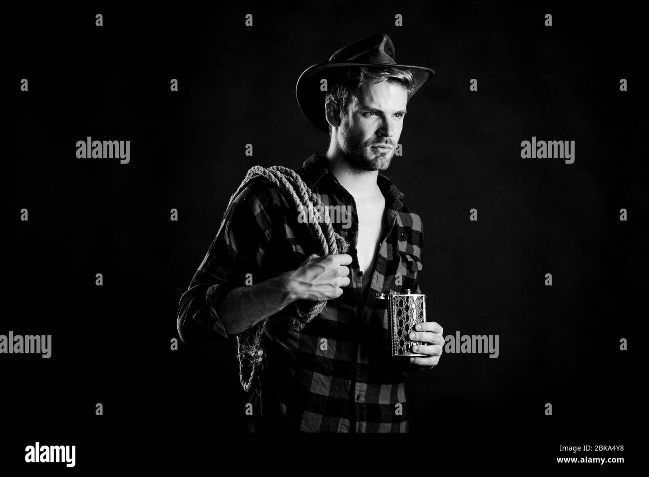 Sheriff concept. Brutal cowboy drinking alcohol. Western culture. Man wearing hat hold rope and flask. Lasso tool of American cowboy. Man handsome unshaven cowboy black background. Western life. Stock Photo