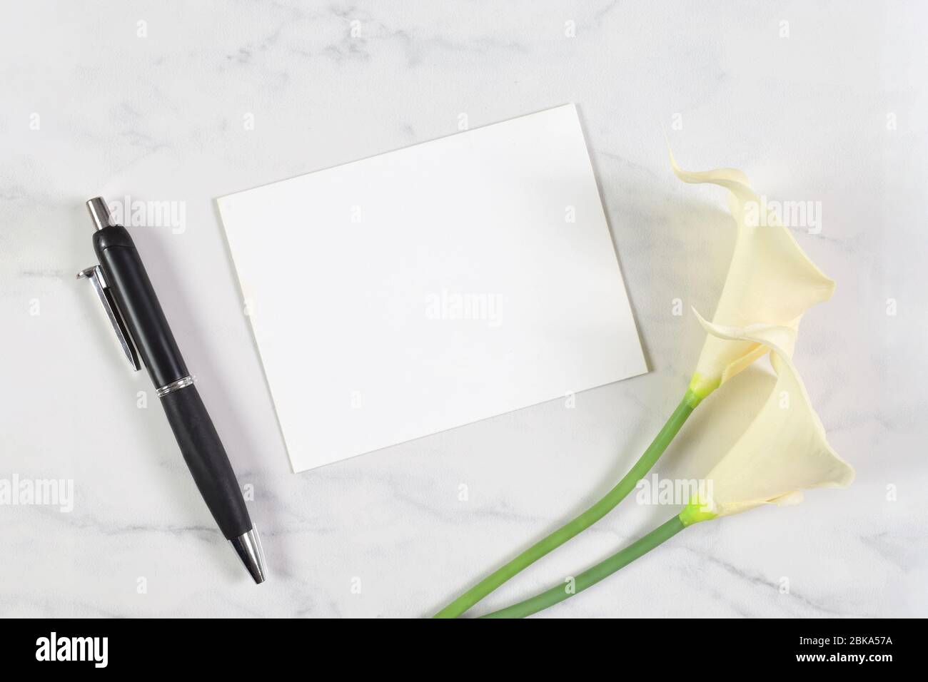 A white card rests atop a white marble background. Calla lilies and a fancy pen surrounded the blank card. Stock Photo