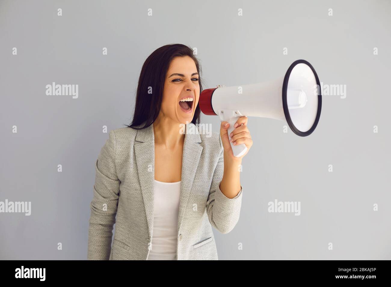 Hispanic woman with a loudspeaker shouts announces news protest on a gray background. Stock Photo