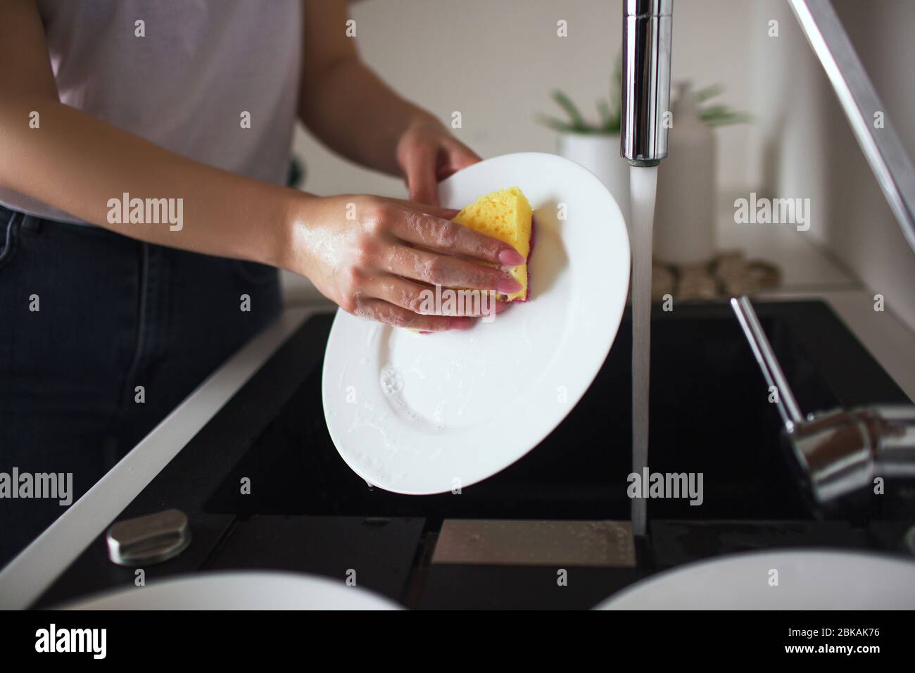 https://c8.alamy.com/comp/2BKAK76/young-woman-in-kitchen-during-quarantine-whshing-white-plates-with-sponge-and-dishwasher-cleaning-dishes-alone-in-kitchen-cut-view-2BKAK76.jpg