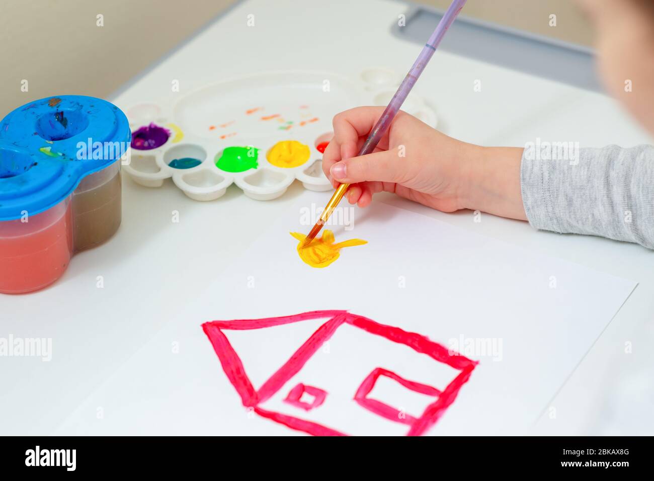 Child is drawing the sun near drawn red house with watercolors on the white sheet of paper. Children creativity. Stock Photo