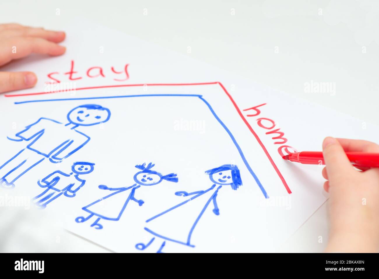 Child is writing words Stay Home with drawn happy family on white sheet of paper at home. Children creativity. Stock Photo