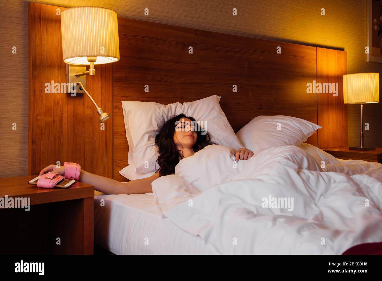 Pretty girl is sleeping in bed with her hand tied to her smartphone with pink handcuffs; she may be an on-call worker or addicted to social media Stock Photo
