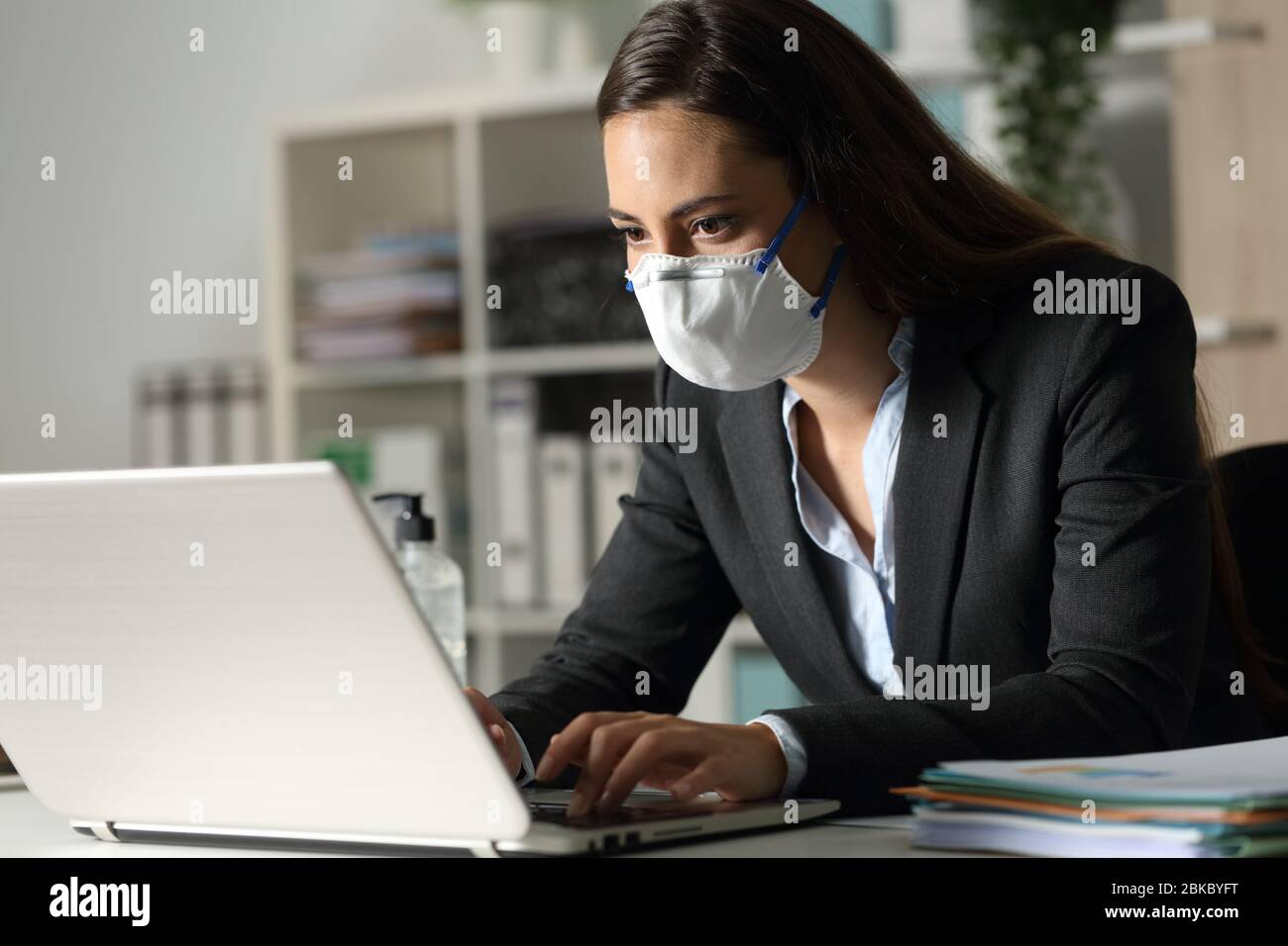 Concentrated executive woman working late on laptop avoiding covid-19 with mask sitting at night in the office Stock Photo