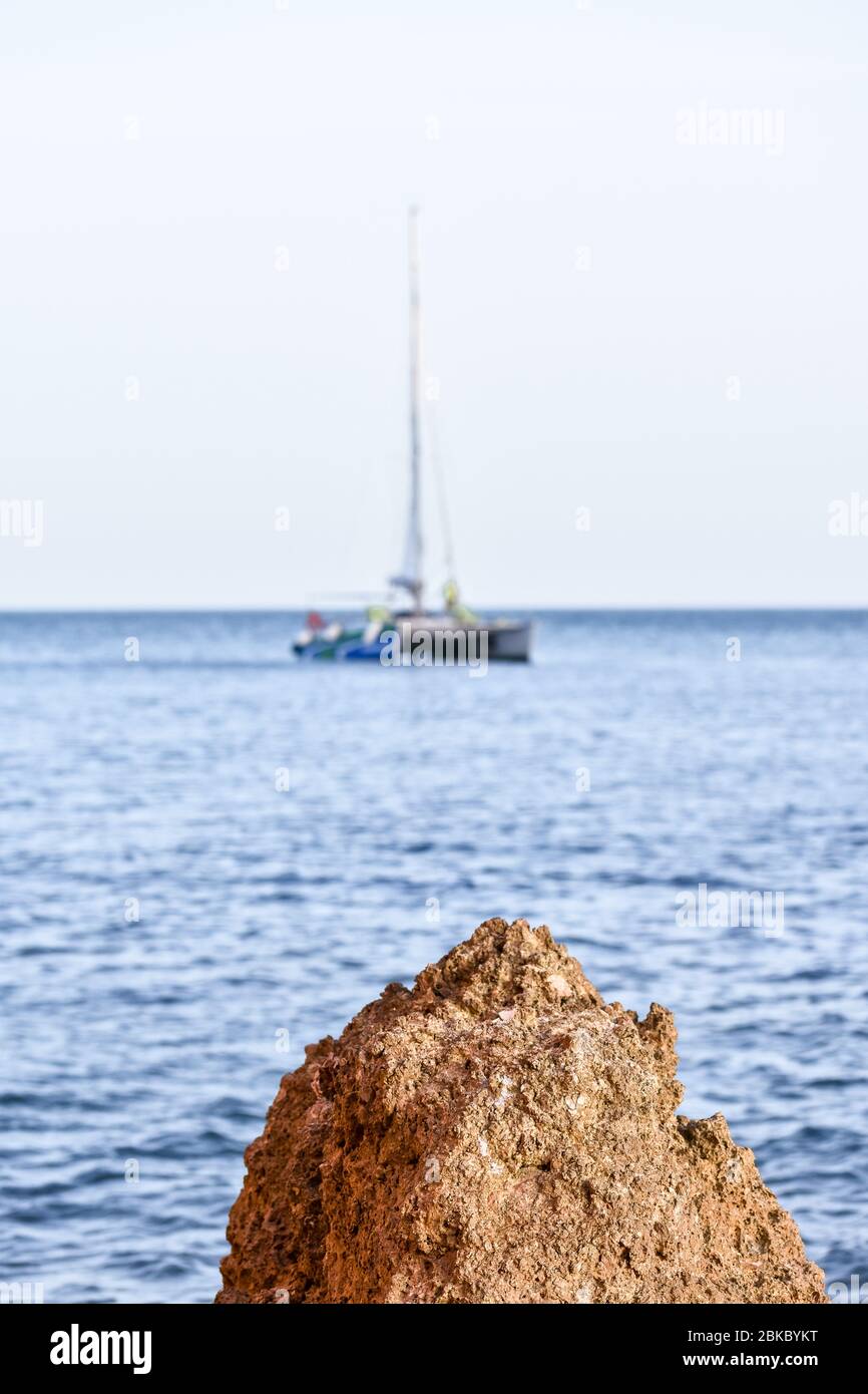 Rock in the foreground, and unfocused sailboat at sea in the background. Concept of sports sailing. Algarve, Portugal Stock Photo