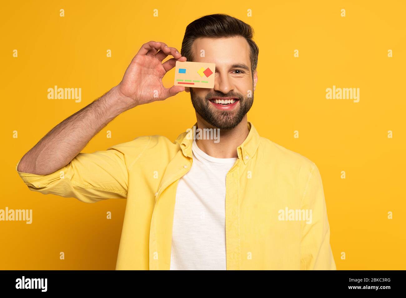 Smiling man covering eye with model of credit card on yellow background ...