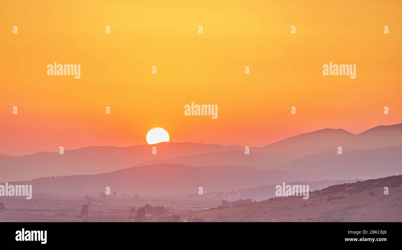 Panorama of beautiful mountains covered with trees at dawn in summer. Bright large orange sun disc. Solar reflection. Flash of the sun. Lens flare. Na Stock Photo