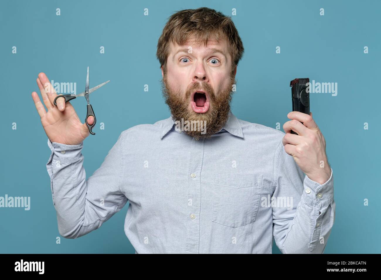 Funny shaggy, bearded man with scissors and a trimmer in hands is shocked because he will have to cut himself during quarantine. Stock Photo