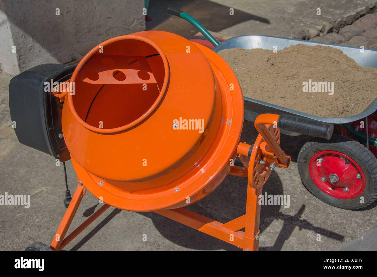 Cement mixer machine is at construction site with wheelbarrow, tools, sand and cement bag. Orange concrete mixer at building construction works Stock Photo