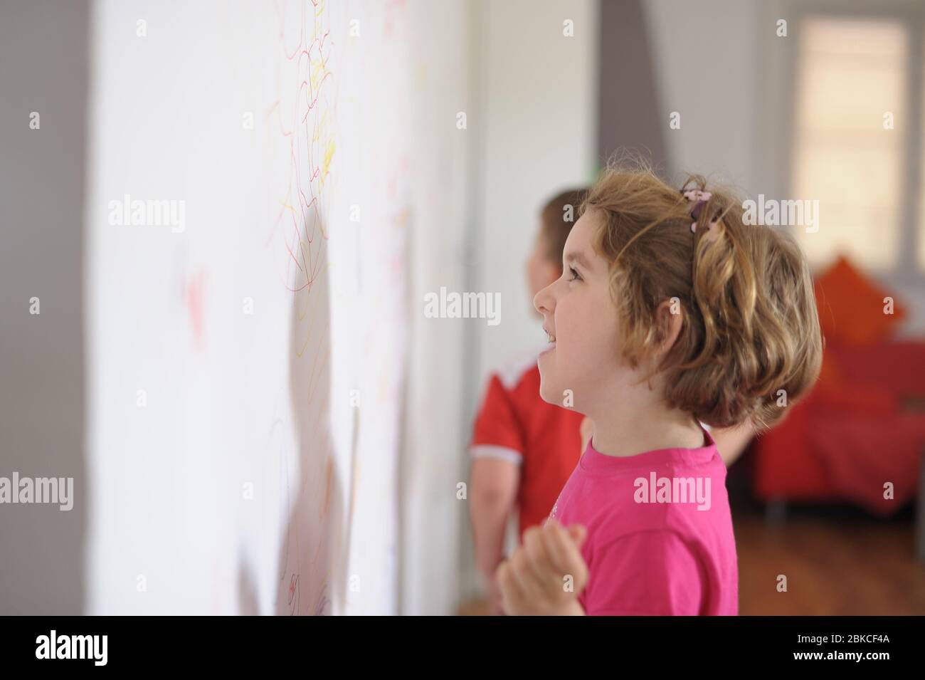 Little blonde girl with short hair drawing with colored pencils on white wall Stock Photo