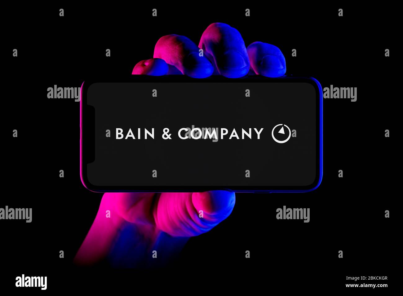 A smartphone displaying the Bains & Company logo is held by a hand shot on a black background (editorial use only). Stock Photo