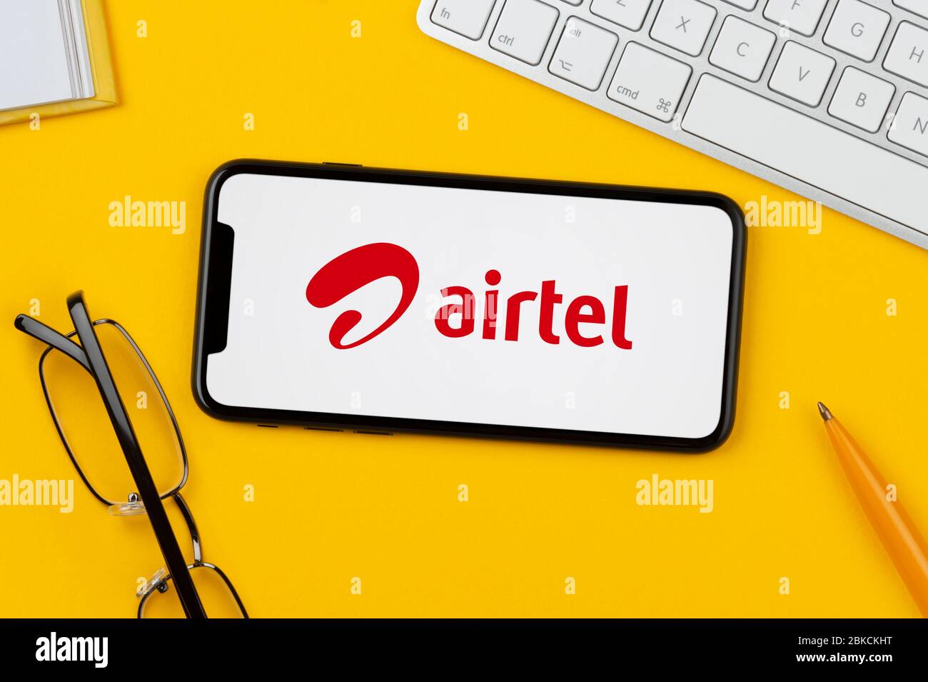 A smartphone showing the Bharti Airtel logo rests on a yellow background along with a keyboard, glasses, pen and book (Editorial use only). Stock Photo