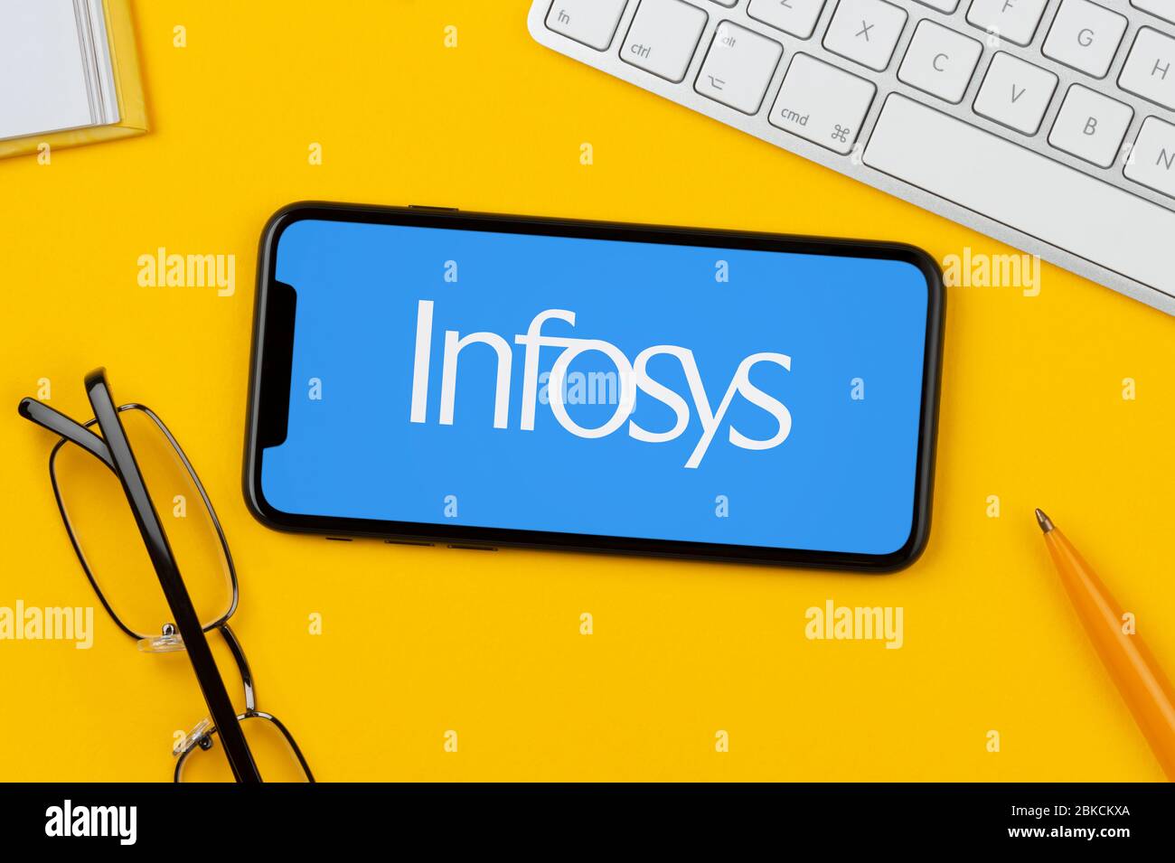 A smartphone showing the Infosys logo rests on a yellow background along with a keyboard, glasses, pen and book (Editorial use only). Stock Photo