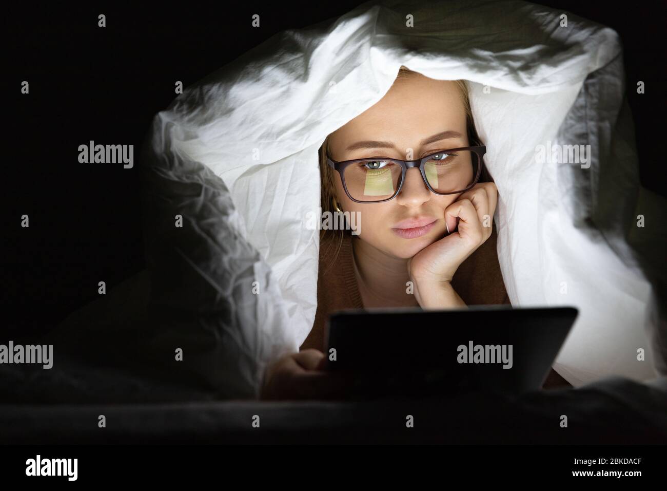 Woman lying in bed under the blanket using tablet late at night, can not sleep, news search about Covid-19 or scrolling social networks. Insomnia, nom Stock Photo