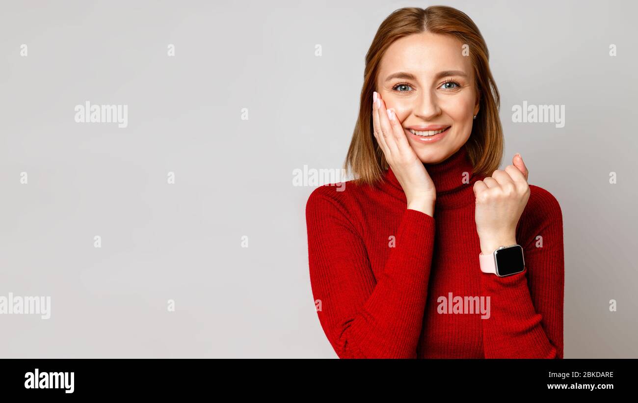Happy stylish business woman in red turtleneck showing smartwatch on wrist, laughing and touching her face by hand, looks on you. Isolated on grey bac Stock Photo