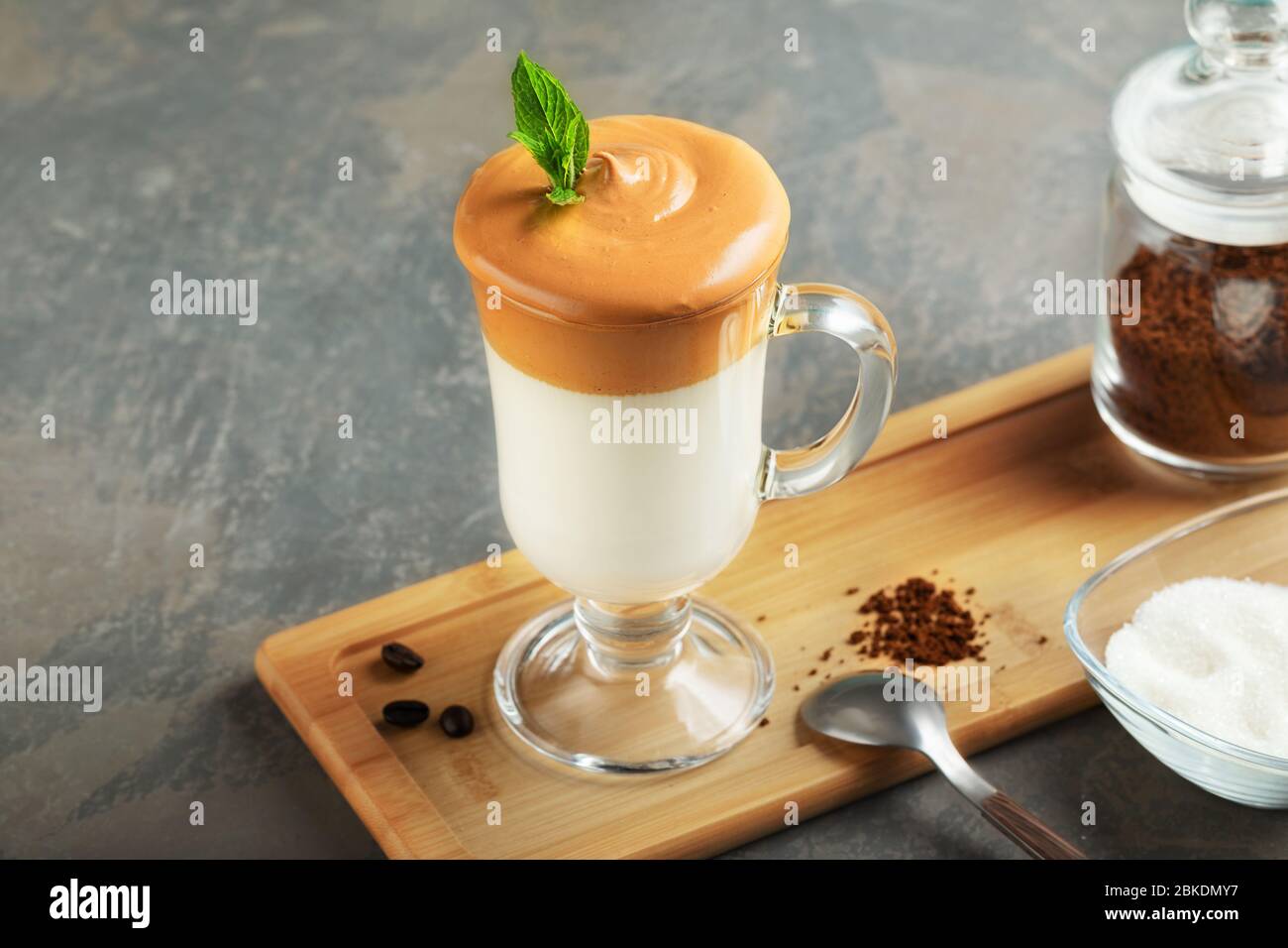 Dalgona frothy coffee in glass on wooden board on grey background. Trend korean Iced  latte coffee drink with foam of instant coffee with ingredients. Stock Photo