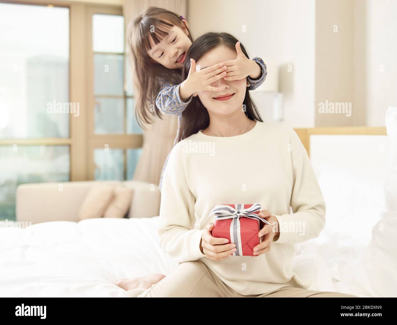 happy little asian girl giving mother a gift and covering mother's eyes with hands Stock Photo