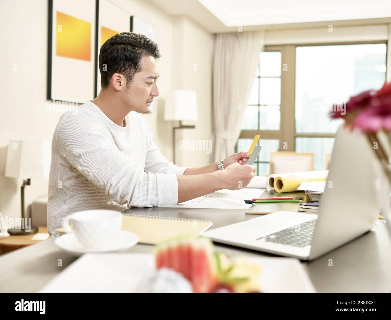 young asian man design professional working from home sitting at kitchen counter comparing two two color cards (artwork in background digitally altere Stock Photo