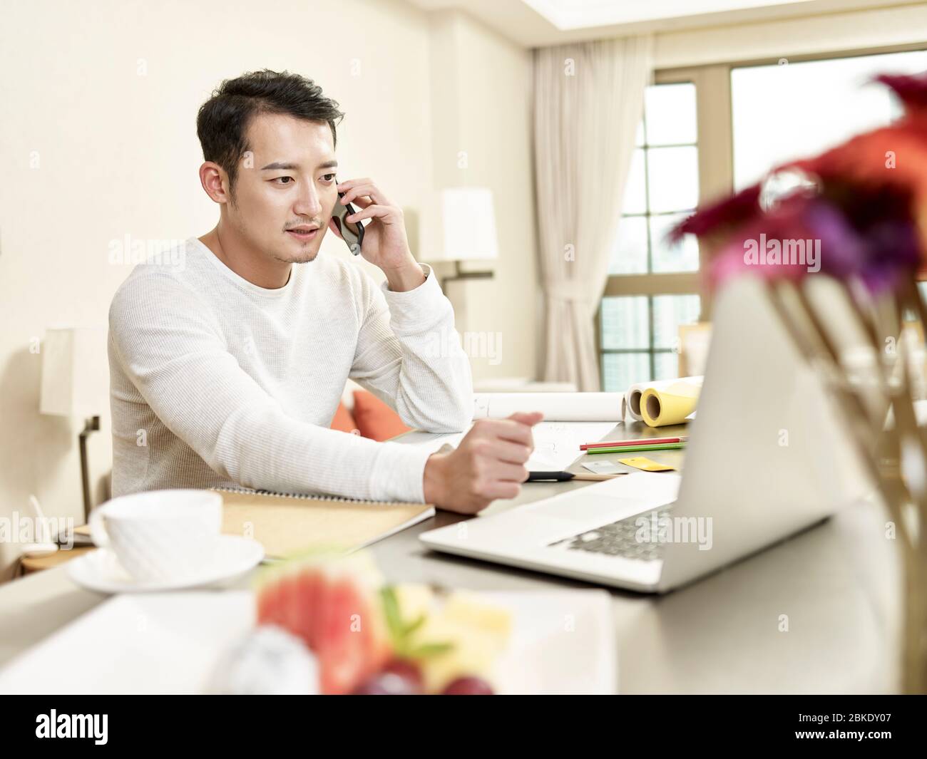 young asian man design professional working from home sitting at kitchen talking on mobile phone Stock Photo