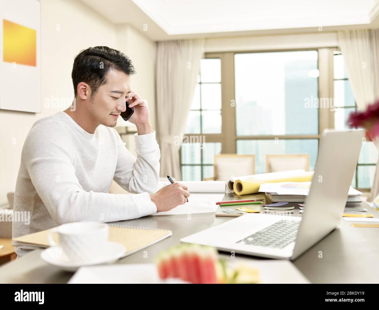 young asian man design professional working from home sitting at kitchen talking on mobile phone (artwork in background digitally altered) Stock Photo