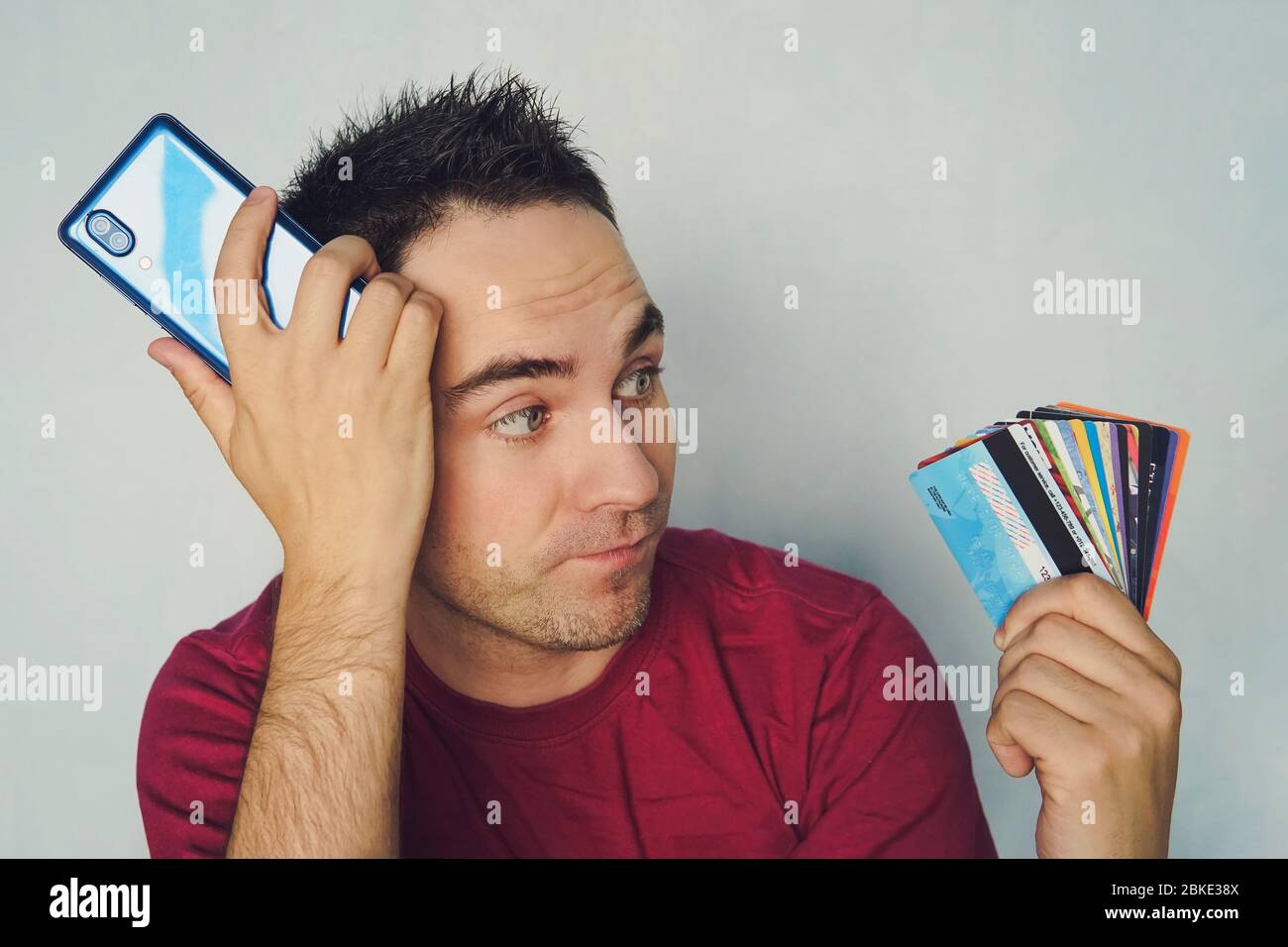 choosing a credit card to pay. Young handsome man thinking about online shopping via internet, wearing red t shirt, standing against blue wall, holdin Stock Photo