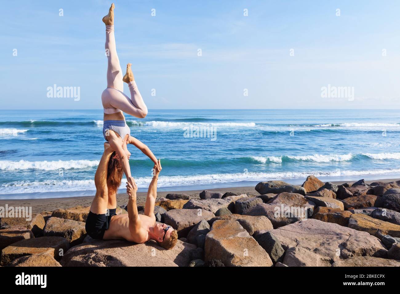 Fit young couple doing acro yoga at spa retreat on sea beach. Active woman on partner feet, balancing at handstand acroyoga pose. Healthy lifestyle. Stock Photo