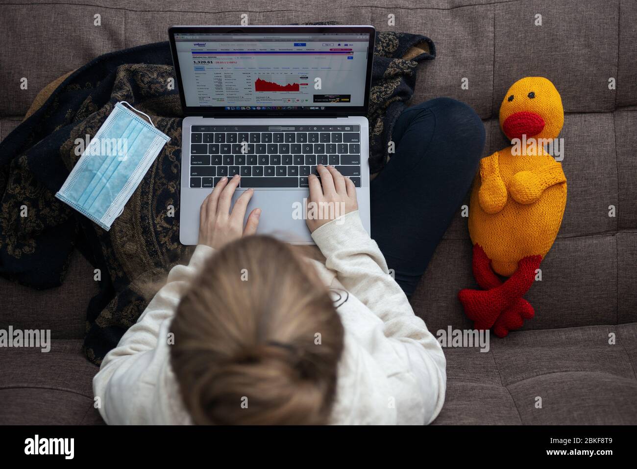 Toronto, Canada - May, 2,2020: Top view of woman sitting on sofa using laptop, with facial mask and stuffed toy. Work from home on the coronavirus qua Stock Photo
