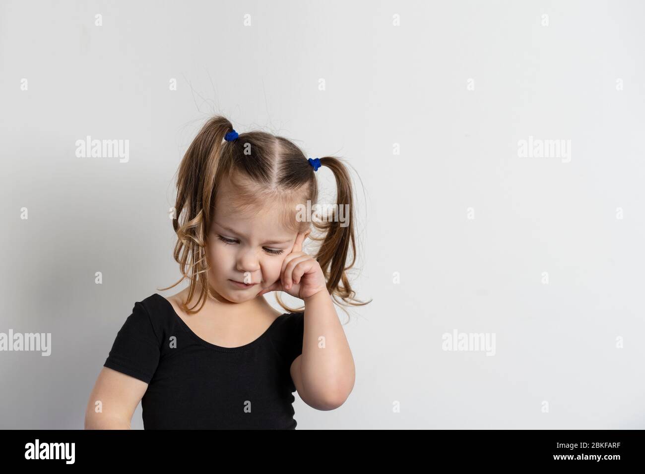 pretty little girl with two ponytails is standing in a pensive pose on a light background. Stock Photo