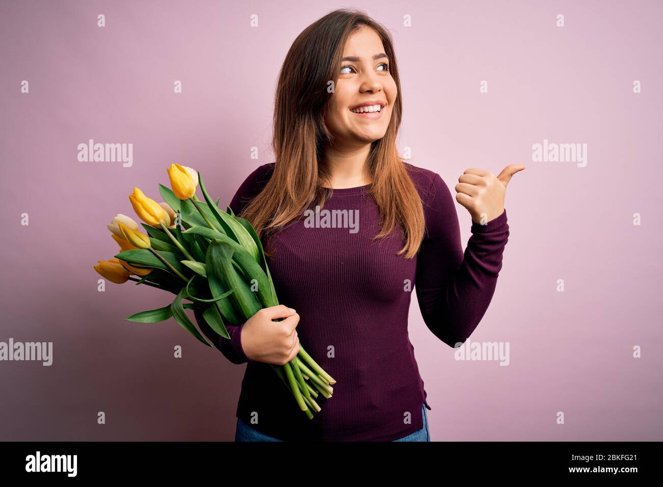 Young blonde woman holding romantic bouquet of yellow tulips flowers over pink background smiling with happy face looking and pointing to the side wit Stock Photo