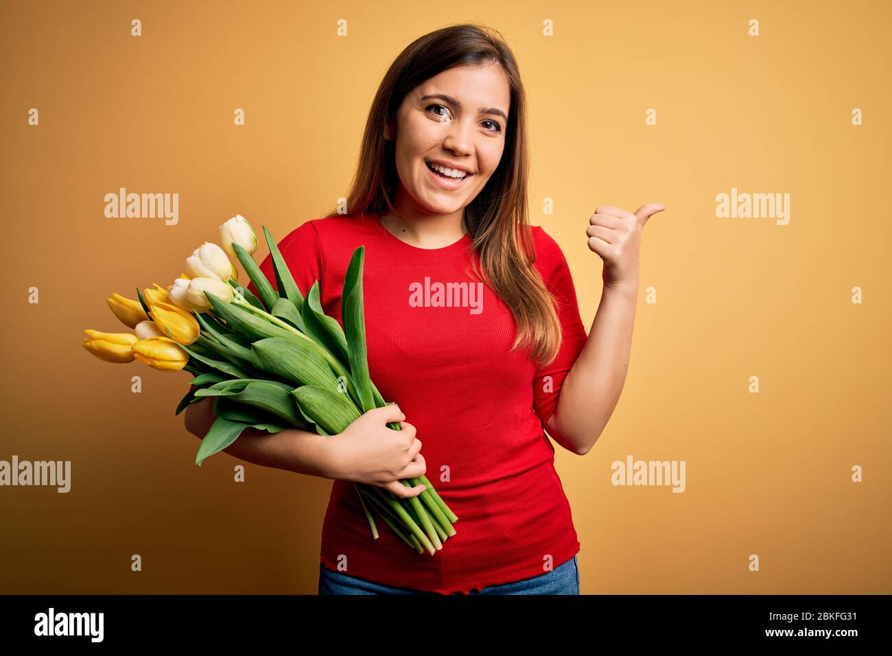 Young blonde woman holding romantic bouquet of tulips flowers over yellow background smiling with happy face looking and pointing to the side with thu Stock Photo