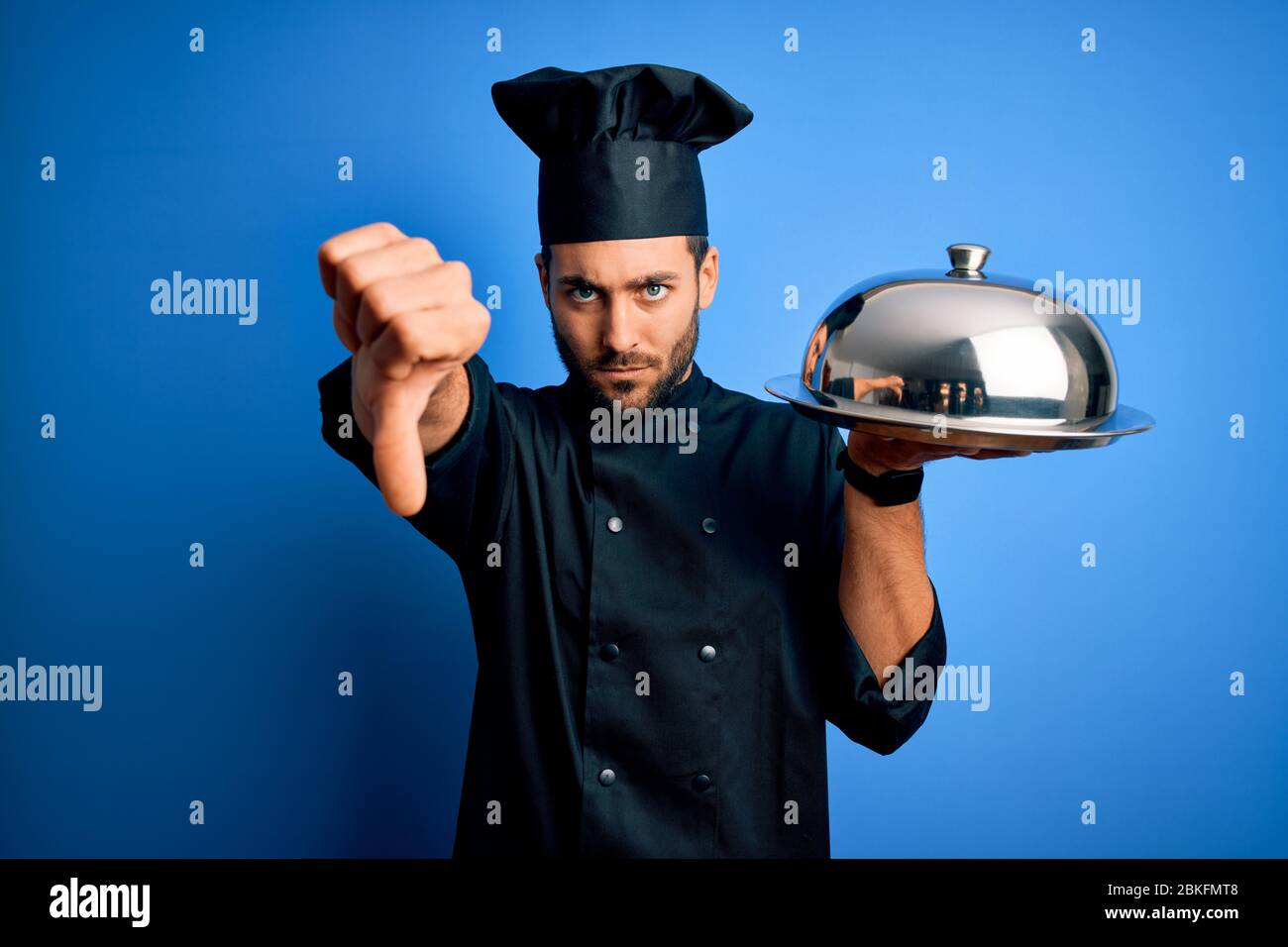 Young cooker man with beard wearing uniform holding tray with dome over ...