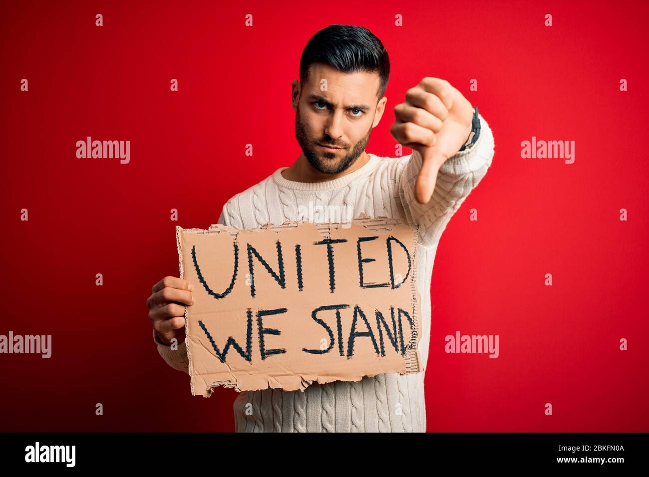 Young handsome activist man protesting holding cardboard with unity message with angry face, negative sign showing dislike with thumbs down, rejection Stock Photo