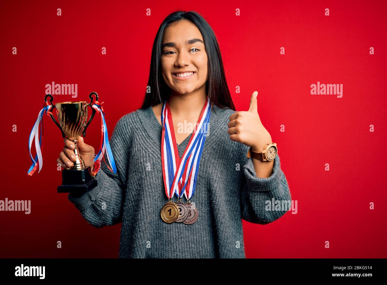 Young beautiful champion asian woman holding trophy wearing medals over ...