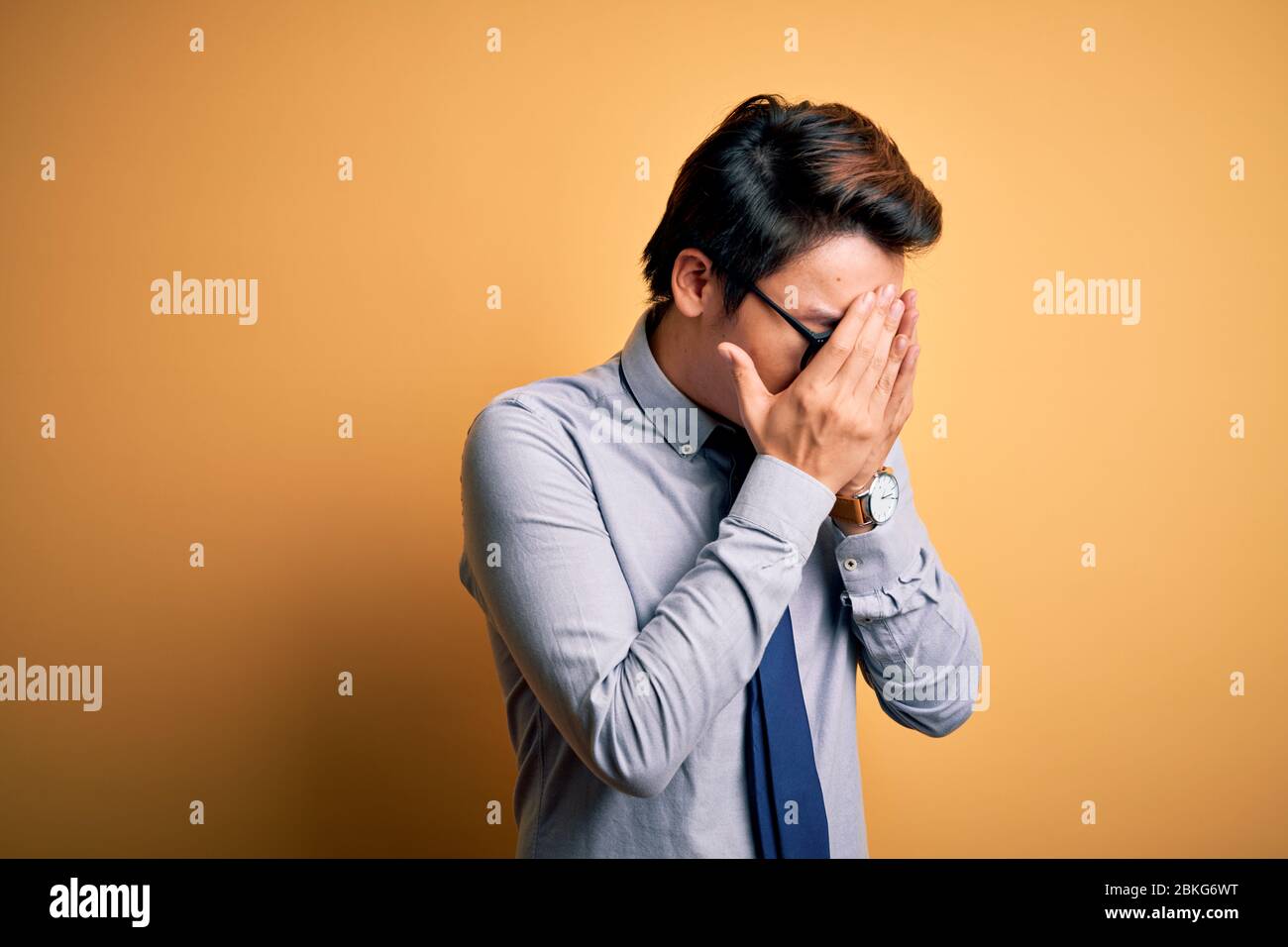 Young handsome chinese businessman wearing glasses and tie over yellow background with sad expression covering face with hands while crying. Depressio Stock Photo