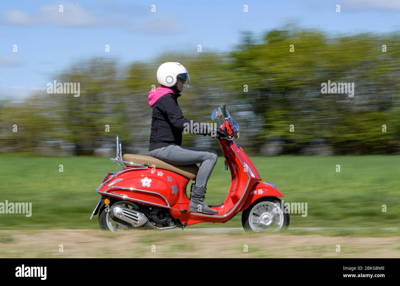 Kiel, Germany. 04th May, 2020. In sunny weather, a woman rides her red 125  cc Vespa scooter along a dirt road wearing safety clothing, helmet and  gloves. Since 01.01.2020, holders of a