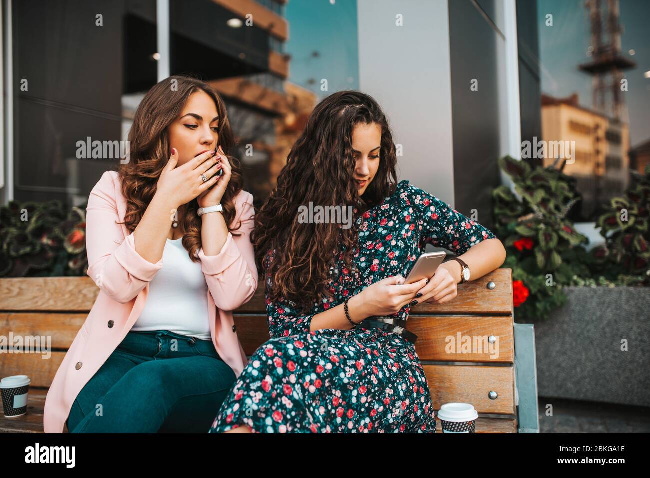 Image of curious woman spying and peeping at smartphone of her friend, while talking on mobile phone and sitting on the bench in the city street Stock Photo