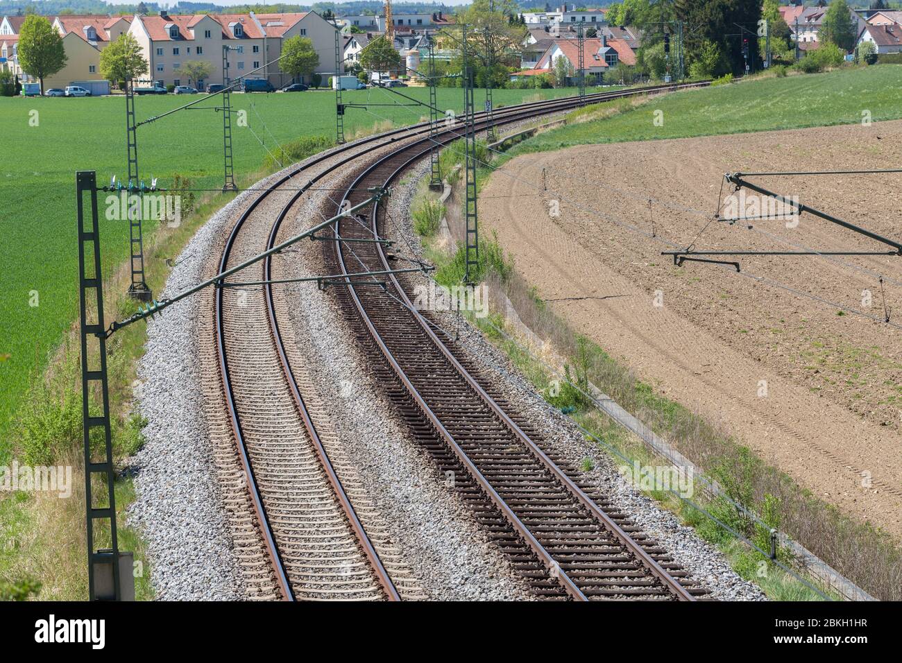 Two railway tracks leading through a landscape with green and brown fields. A town on the horizon. With bend / curve. Symbol for journey, travel. Stock Photo