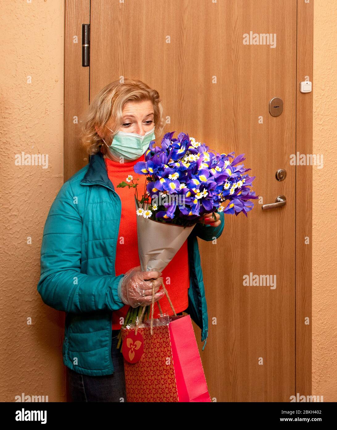 Courier in protective mask and in medical gloves delivers flowers and gift to senior customer on birthday. Delivery service under quarantine,disease o Stock Photo